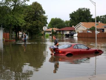 Picture of man sitting on top of car in the middle of flooded area.